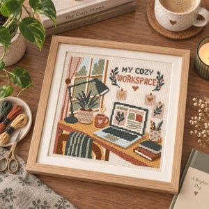 May include: A framed cross-stitch embroidery featuring a desk scene with a laptop, a lamp, a potted plant, and a mug. The text "MY COZY WORKSPACE" is stitched at the top. The design includes a window, books, and wall art, rendered in a palette of muted earth tones, greens, and browns within a light wood frame.