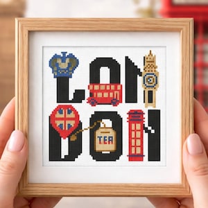 May include: A framed cross-stitch artwork spelling "LONDON" with iconic London imagery. The design includes a crown, red double-decker bus, Big Ben clock tower, a red telephone box, and a tea bag. The frame is made of light-colored wood.
