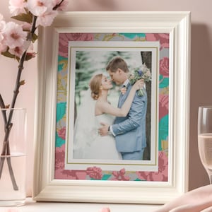 Könnte beinhalten: Weiß gerahmtes Foto eines sich umarmenden Paares mit floralem Rand in Rosa, Türkis und Gold. Der Rahmen steht auf einem Tisch mit einem Glas Champagner und einer Vase mit rosa Blumen.