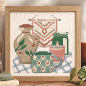 May include: Cross-stitch artwork in a wooden frame. The artwork depicts a still life with a vase, mug, candle, and decorative banner. The color palette includes shades of green, brown, and pink. The mug has a diamond and heart pattern. The artwork is on a blue and white checkered cloth.