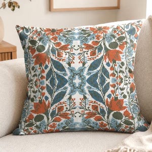 May include: A square throw cushion featuring a symmetrical cross-stitch pattern. The design displays a repeating floral motif in shades of burnt orange, muted blue, and dark green against a white background, creating a balanced, intricate visual.