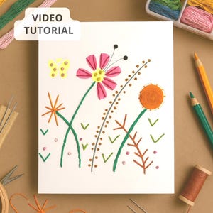 May include: A white card featuring a colorful floral embroidery design with pink, orange, and green thread. The text "VIDEO TUTORIAL" is in a white box. Embroidery supplies, including thread, needles, and pencils, are arranged around the card.