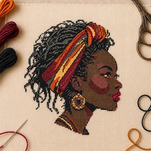 May include: A cross-stitch embroidery on cream fabric depicting the profile of a person with dark skin, black hair, and a red and orange headwrap. The person wears a circular patterned earring and a matching necklace, surrounded by embroidery floss.