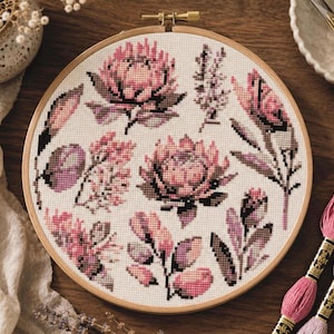 May include: A circular wooden embroidery hoop containing a cross-stitch pattern of various pink and purple protea flowers and botanical sprigs on white Aida fabric. The design features multiple floral clusters with detailed shading and textures.