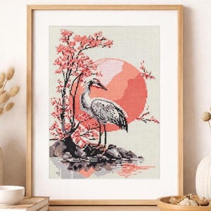 May include: Cross-stitch artwork in a wooden frame, depicting a crane by water with a coral sun and cherry blossom tree. The image uses a palette of grey, coral, and white. The artwork evokes a sense of calm and is inspired by nature. The frame is made of wood.