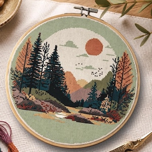 May include: A completed cross-stitch landscape scene in a wooden embroidery hoop. The design features a forest with evergreen trees, mountains, a setting sun, and a river. The colour palette includes greens, browns, oranges, and blues.