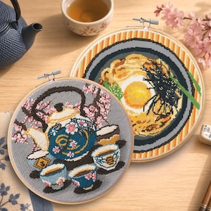 May include: Two cross-stitch embroidery hoops featuring Japanese themes. One depicts a blue teapot, cups, and cherry blossoms. The other shows a bowl of ramen with an egg. A blue teapot and tea cup are in the background.