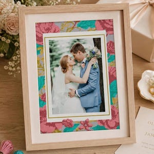 May include: A light wood picture frame containing a photograph of a couple in wedding attire. The photo is surrounded by a cross-stitch border featuring a floral pattern in shades of pink, teal, and yellow on a gray background.