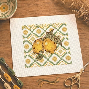 May include: A completed cross-stitch project featuring two yellow lemons with green leaves, set against a geometric pattern in yellow and green. The needlework is on white fabric, with spools of thread and scissors nearby.