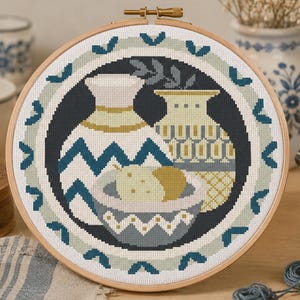 Könnte beinhalten: Fertige Kreuzstich-Stickerei in einem Holzreifen. Das Design zeigt zwei Vasen und eine Schale mit Obst in Blau-, Grau-, Gelb- und Weißtönen. Die Stickerei ist von einem dekorativen Rand umgeben.