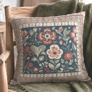 Op de afbeelding: Een vierkant sierkussen met een bloemenpatroon in kruissteekstijl op een donkerblauwe achtergrond. Het ontwerp bevat rode, witte en saliegroene bloemen en bladeren, omlijst door een decoratieve rand met geometrische motieven in bijpassende kleuren.