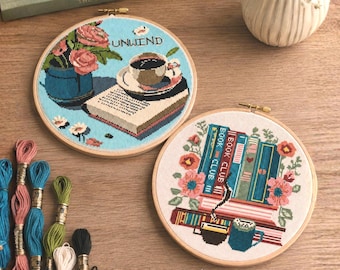 May include: Two circular wooden embroidery hoops featuring cross-stitch designs. One hoop shows a blue background with a book, a cup of coffee, and pink flowers with the text UNWIND. The other hoop displays a stack of books labeled BOOK CLUB.