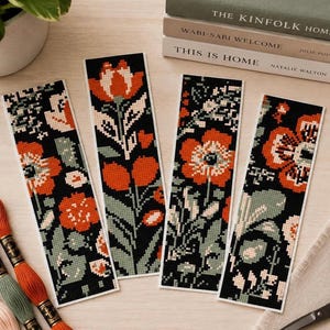 May include: Four rectangular bookmarks featuring cross-stitch floral patterns in shades of orange, sage green, and cream on a black background. The designs include various flower and leaf motifs arranged in vertical compositions on each bookmark.