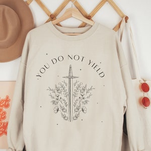 May include: A beige sweatshirt with a black graphic of a sword surrounded by flowers and the text "You Do Not Yield".