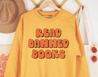 May include: Yellow sweatshirt with the text "READ BANNED BOOKS" in orange letters. The letters are in a retro, bubble-like font.