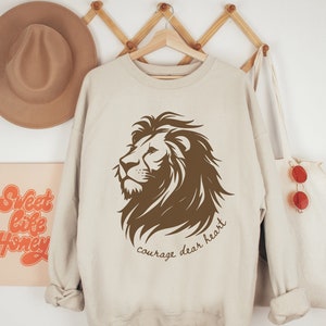 May include: A beige sweatshirt with a brown lion graphic and the text "courage dear heart".