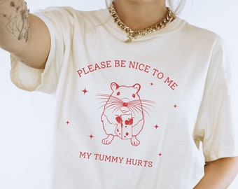 May include: A white t-shirt with a red graphic of a hamster holding a piece of cheese. The text "Please be nice to me" and "My tummy hurts" is printed on the shirt.