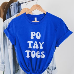 May include: A royal blue t-shirt with white text that reads "PO TAY TOES". The shirt is hanging on a hanger with a denim shirt and a brown hat in the background.