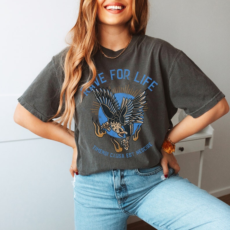May include: A black t-shirt with a graphic of a blue eagle in flight with flames on either side. The text 'LIVE FOR LIFE' is written in a circle around the eagle. The text 'TIMENDI CAUSA EST NESCIRE' is written below the eagle.