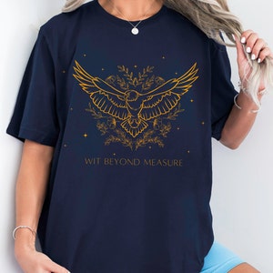 May include: Navy blue t-shirt with a gold outline of a bird with wings spread, surrounded by flowers and stars. The text "WIT BEYOND MEASURE" is printed below the bird.