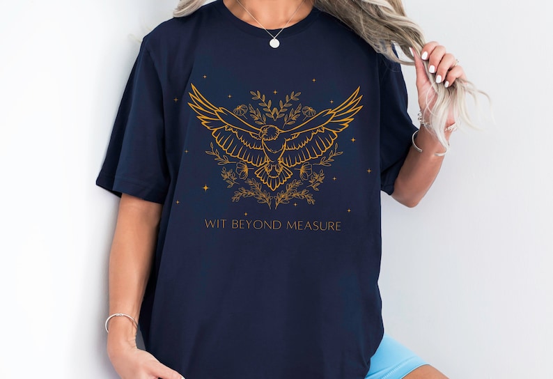 May include: Navy blue t-shirt with a gold outline of a bird with wings spread, surrounded by flowers and stars. The text "WIT BEYOND MEASURE" is printed below the bird.
