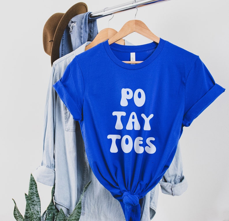 May include: A royal blue t-shirt with white text that reads "PO TAY TOES". The shirt is hanging on a hanger with a denim shirt and a brown hat in the background.