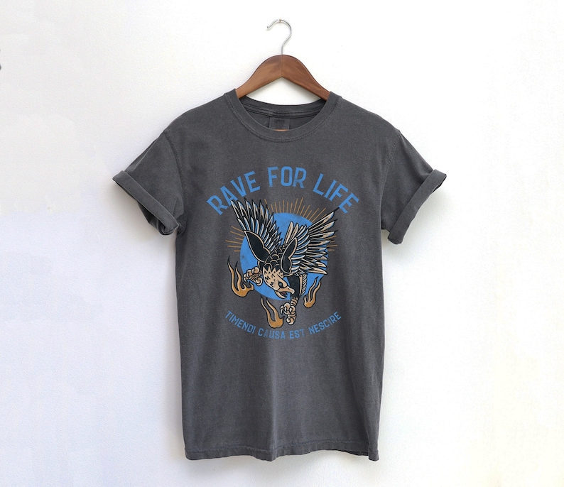 May include: A dark gray t-shirt with a graphic of a flying eagle with the text "Rave For Life" and "Trendi Canga Est Nescore" printed on the front.