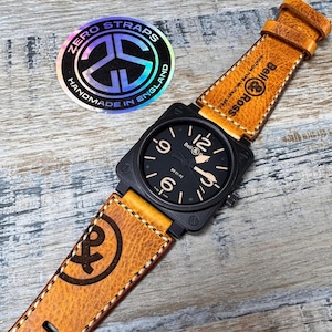 Puede incluir: Una correa de reloj de cuero marrón con costuras blancas y una esfera de reloj Bell & Ross negra. La correa tiene el texto "ZERO STRAPS" y "HANDMADE IN ENGLAND" en una pegatina. La correa tiene un logotipo "Bell & Ross" y el texto "AVIATION TYPE-MILITARY SPEC" en la correa.