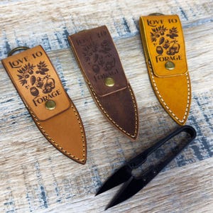 May include: Three leather keychains with the text "Love to Forage" and a design of a tree with leaves and berries. The keychains are brown, tan, and yellow. A small black pair of scissors is also in the image.