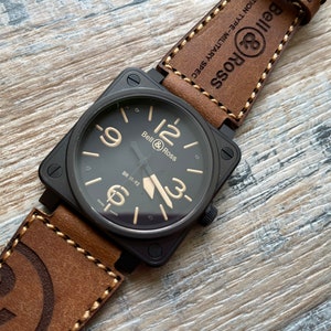 May include: A black Bell & Ross BR 31-92 watch with a square face and tan numerals. The watch has a brown leather strap with white stitching and the text "Bell & Ross" and "MILITARY SPEC".
