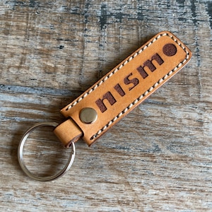 May include: A brown leather keychain with a white stitched border and the word "nismo" embossed in brown lettering.