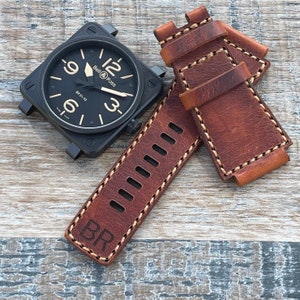 May include: A brown leather watch strap with white stitching and the letters "BR" embossed on the strap. The strap is designed for a square watch with a black face and white numbers.