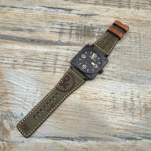 May include: A black square watch with a green canvas strap and brown leather detailing. The watch face is black with white numerals. The strap features the text "Bell & Ross" and a logo. The watch is on a wooden surface.