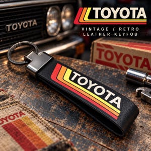Pode incluir: Um porta-chaves de couro preto com um anel de metal e um gráfico impresso com a palavra TOYOTA em branco, acompanhado por três faixas horizontais em vermelho, laranja e amarelo. O design inclui o texto VINTAGE / RETRO LEATHER KEYFOB.
