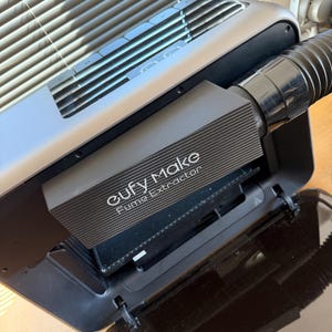 Könnte beinhalten: Nahaufnahme eines schwarzen und grauen Eufy Make Fume Extractor. Das Gerät hat einen rechteckigen Körper mit strukturierter Oberfläche und einem flexiblen schwarzen Schlauch. Der Text "Eufy Make Fume Extractor" ist auf der Vorderseite aufgedruckt.