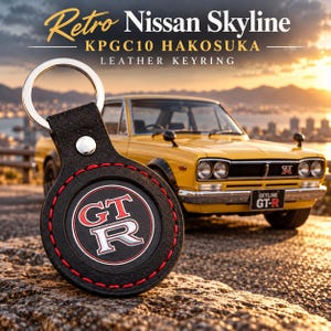 May include: A round black leather keyring featuring a silver metal ring and red stitching. The center displays a circular emblem with the red and white text GT-R. The text Retro Nissan Skyline KPGC10 HAKOSUKA LEATHER KEYRING is visible.