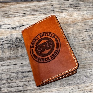 May include: A tan leather wallet displaying the Royal Enfield logo. The circular emblem reads "ROYAL ENFIELD SINCE 1901". The wallet has white stitching around the edges and a smooth, textured surface.
