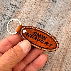 Puede incluir: Llavero de cuero marrón con el texto "BMW R1200RT" en relieve. El llavero de forma ovalada tiene un llavero plateado y costuras blancas alrededor de los bordes. Un remache plateado asegura el cuero al llavero.