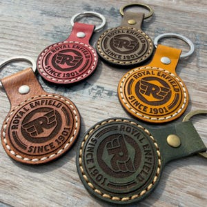 May include: A collection of leather keychains in various colors, including red, brown, and green. Each keychain features the "ROYAL ENFIELD SINCE 1901" logo, with a metal ring and a leather strap for attachment.