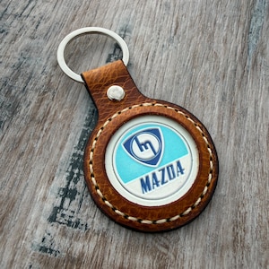 Op de afbeelding: Een ronde bruin lederen sleutelhanger met een zilverkleurige metalen ring en een centraal rond embleem. Het embleem toont een blauw-wit schildlogo met de tekst MAZDA in blauwe hoofdletters op een witte achtergrond onder het schild.