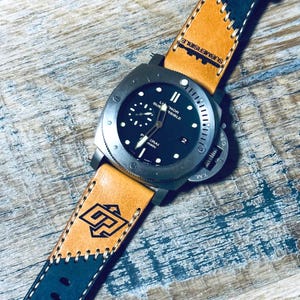 May include: A black-faced watch with a stainless steel case and a tan and navy leather strap. The watch face has white hands and markers, with the text "LUMINOR SUBMERSIBLE" and "PANERAI" visible. The strap features a stitched design and a logo.