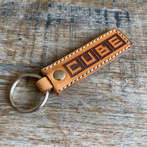 Può includere: Un portachiavi in ​​cuoio color beige con un bordo cucito marrone e un anello portachiavi argentato. La parola "CUBE" è impressa in marrone sul cuoio.