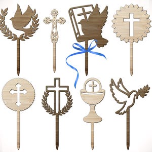May include: A collection of wooden cake toppers featuring religious symbols. Designs include crosses, doves, and a chalice. Some toppers have decorative elements like laurel wreaths and a blue ribbon. The toppers are made of light brown wood.