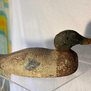 May include: A vintage wooden duck decoy with a weathered appearance. The duck has a green head, a brown body with a blue accent, and a yellow beak. The decoy is displayed on a clear acrylic surface.