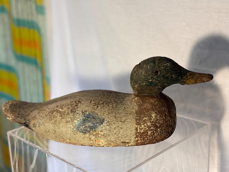 May include: A vintage wooden duck decoy with a weathered appearance. The duck has a green head, a brown body with a blue accent, and a yellow beak. The decoy is displayed on a clear acrylic surface.