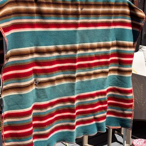 May include: A rectangular blanket featuring horizontal stripes in shades of teal, red, brown, and cream. The fabric displays a visible small hole on the left side. The pattern consists of repeating bands of varying widths across the entire surface.