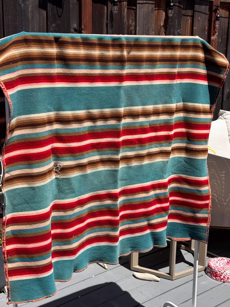May include: A rectangular blanket featuring horizontal stripes in shades of teal, red, brown, and cream. The fabric displays a visible small hole on the left side. The pattern consists of repeating bands of varying widths across the entire surface.