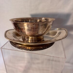 May include: A silver-colored bowl and saucer set. The bowl sits atop a pedestal base, resting on a matching saucer. The rim of the saucer has a decorative rope-like border. The set is displayed on a clear acrylic stand.