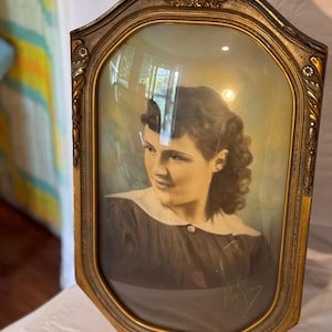 May include: An antique portrait in a gold-toned, ornate frame. The oval-shaped image features a woman in a dark dress with a white collar. The frame has decorative scrollwork at the top and a signature in the bottom right corner.