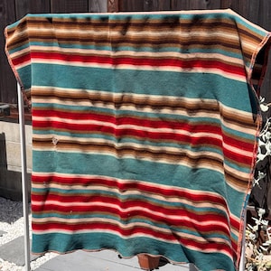 May include: A rectangular woven blanket featuring horizontal stripes in shades of teal, red, tan, and dark brown. The fabric displays a consistent pattern of varying widths across the entire surface, with a small visible tear on the left side.